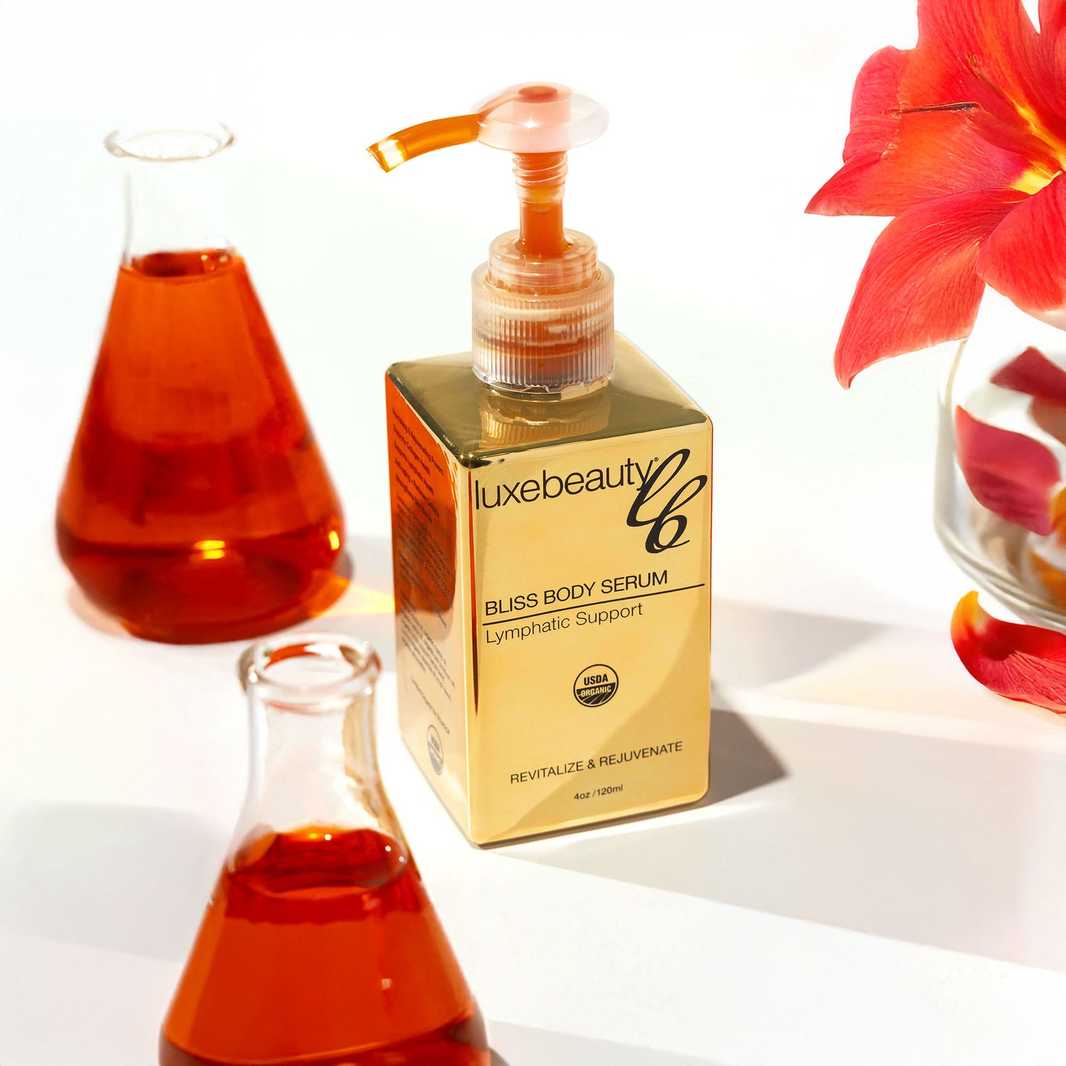 Gold bottle of luxebeauty Bliss Body Serum with red liquid bottles and a flower in the background.